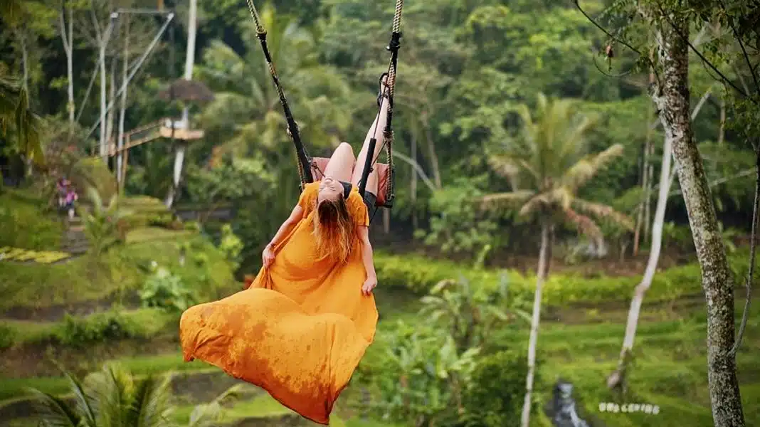 A person in a bright orange dress swings high above the lush Ubud rice fields, a moment of joy and freedom.